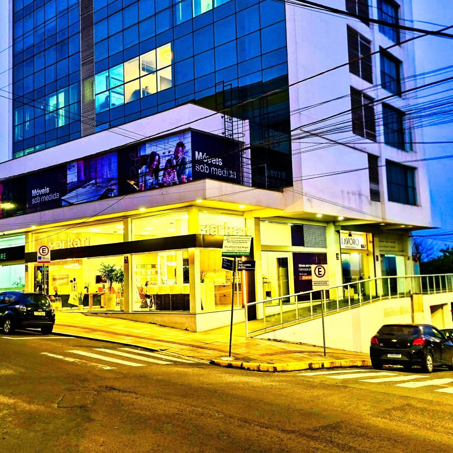 Fachada de edifício comercial ao anoitecer, com lojas iluminadas no térreo e fachada envidraçada