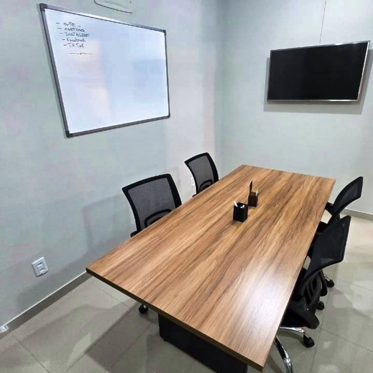 Sala de reuniões com mesa de madeira, cadeiras pretas, quadro branco e televisor na parede